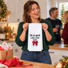 Woman holding a white kitchen towel with I'm So Good Santa Came Twice lettering and red Christmas bow at a holiday party